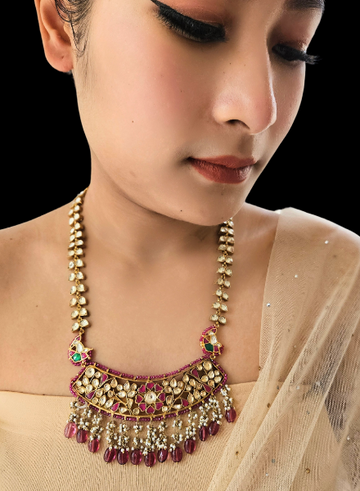 A woman wearing a traditional kundan and ruby necklace with white lusters, featuring a kundan chain and ruby drops.
