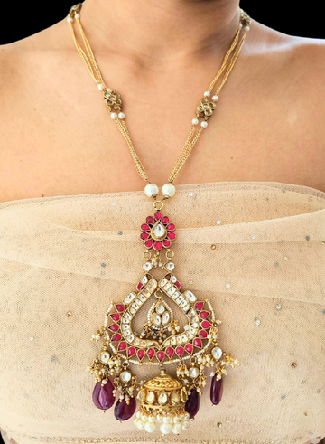 A person wearing a gold-colored necklace with a ruby and kundan pendant, accented with a gold and kundan jhumki, and a double gold chain.