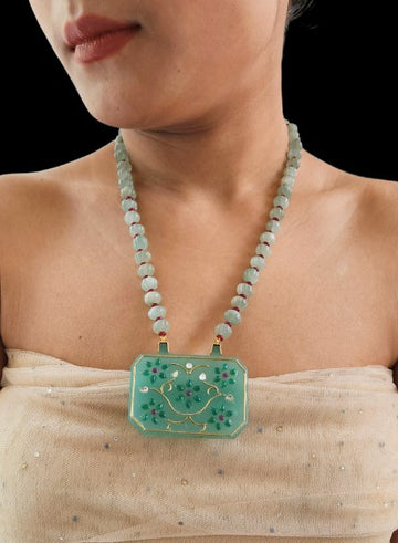A person wearing a geometric green onyx pendant necklace with gold and stone inlay, accompanied by a string of aventurine stones.